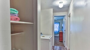 Un couloir étroit avec un mur blanc, un plancher en bois et un luminaire au plafond. Une porte donne sur une pièce où se trouvent un lit et une fenêtre ornée d’un rideau bleu.