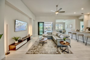 A modern living room with a sofa, coffee table, and TV. There is a kitchen area with bar stools and a dining table. - Villa Bright Side - on water, pool, spa, modern (Cape Coral)