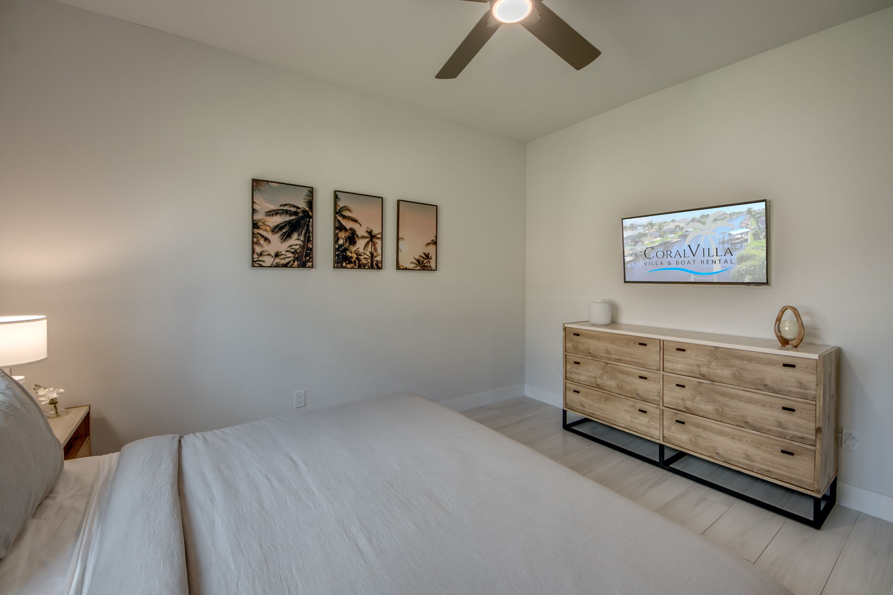 Een slaapkamer met een bed, nachtkastje, dressoir en plafondventilator. Er hangen drie foto’s aan de muur en een televisie waarop “CoralVilla Villa & Boat Rental” staat.