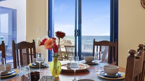 Mesa de comedor lista con platos, tazones, vasos y un jarrón con flores, con vista a una playa.