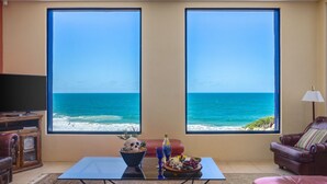 Una sala de estar con un amplio ventanal con vistas al océano, un televisor, una mesa de centro con objetos decorativos y un sillón de cuero.
