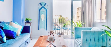 A modern living room with a blue sofa, a wooden coffee table, and a blue armchair. There is a balcony with a view of buildings and plants.
