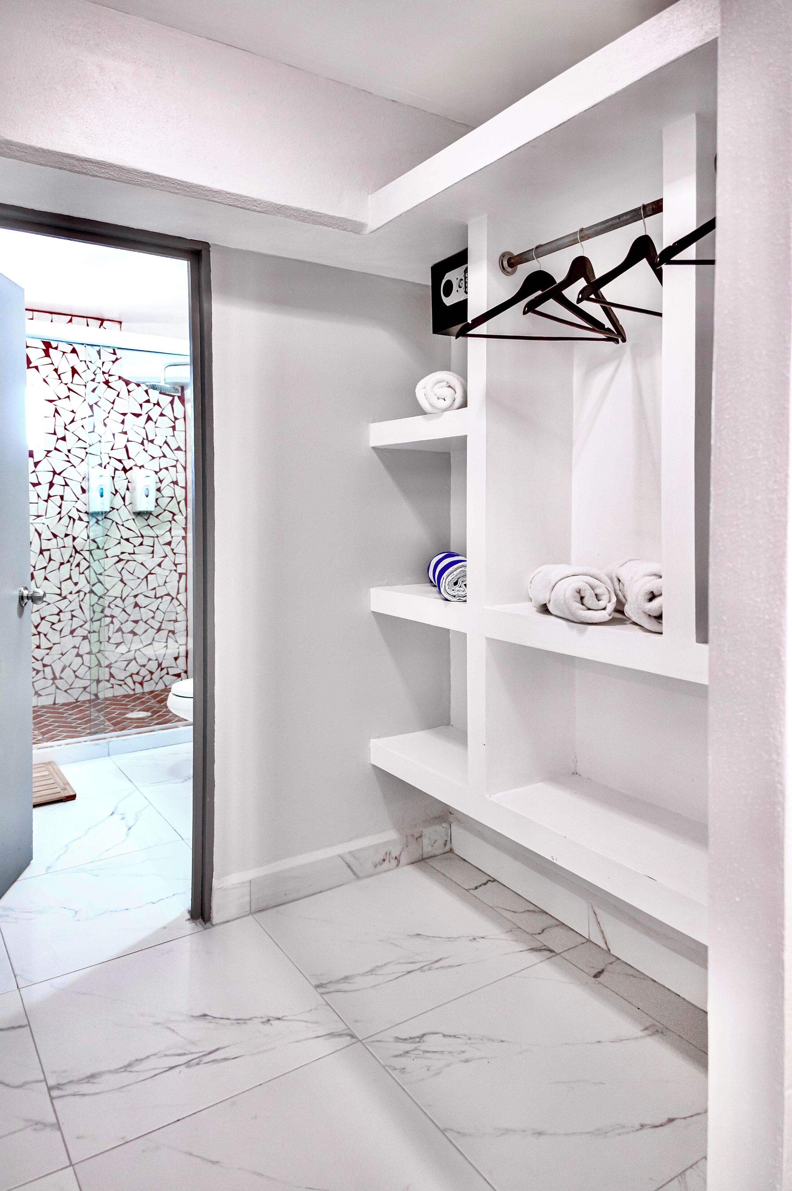 A bathroom with white tiled floors, a shower area with a mosaic wall, and a white shelving unit with towels and a striped towel.