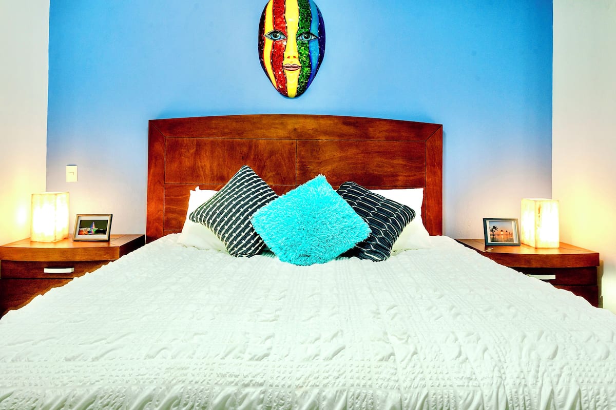 Un letto rifatto con cura, con una testiera in legno, due comodini con lampade e un quadro colorato sopra il letto.