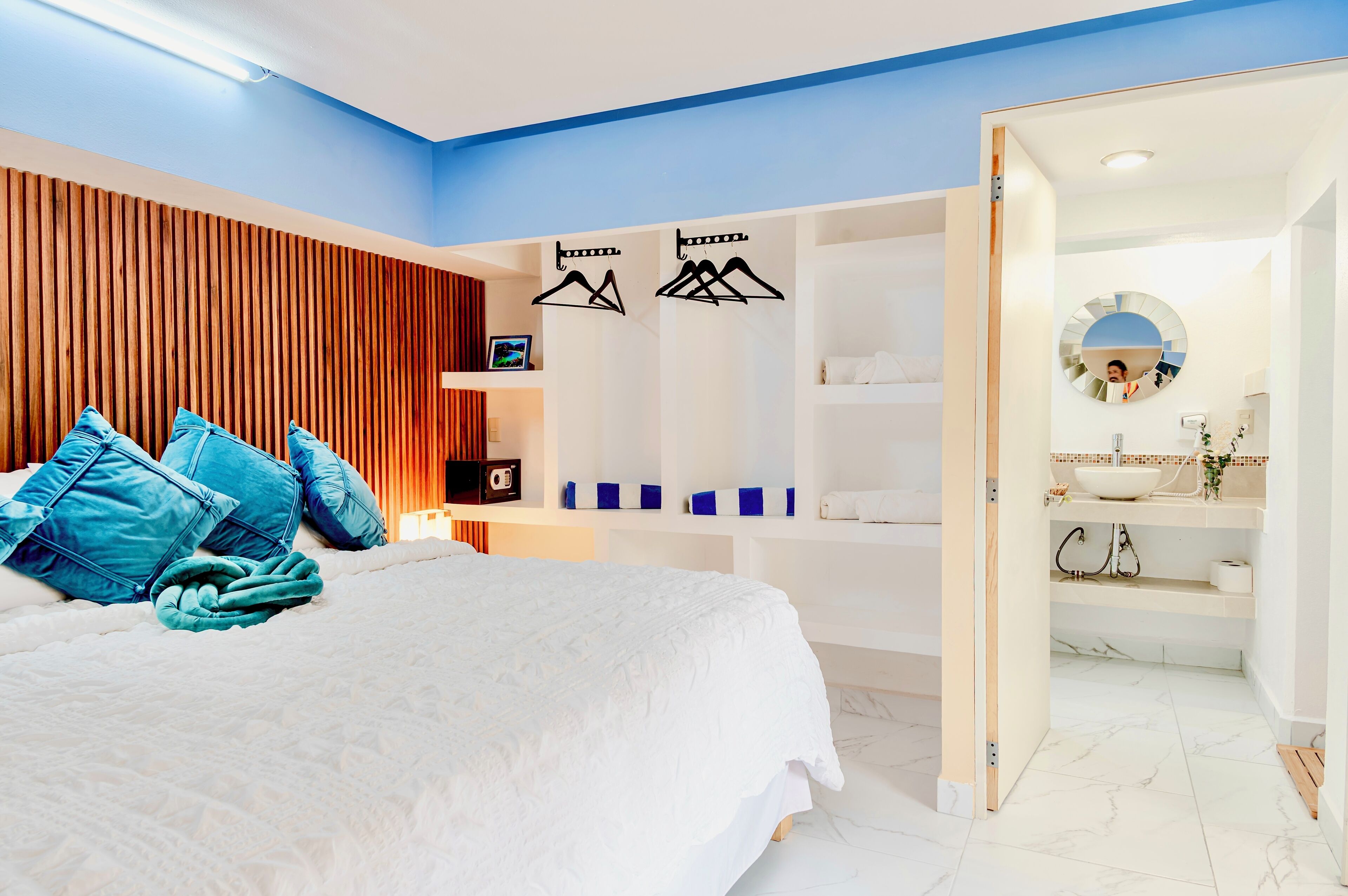 A modern bedroom with a large bed, blue pillows, a wooden headboard, a wardrobe, and a bathroom with a mirror and sink.