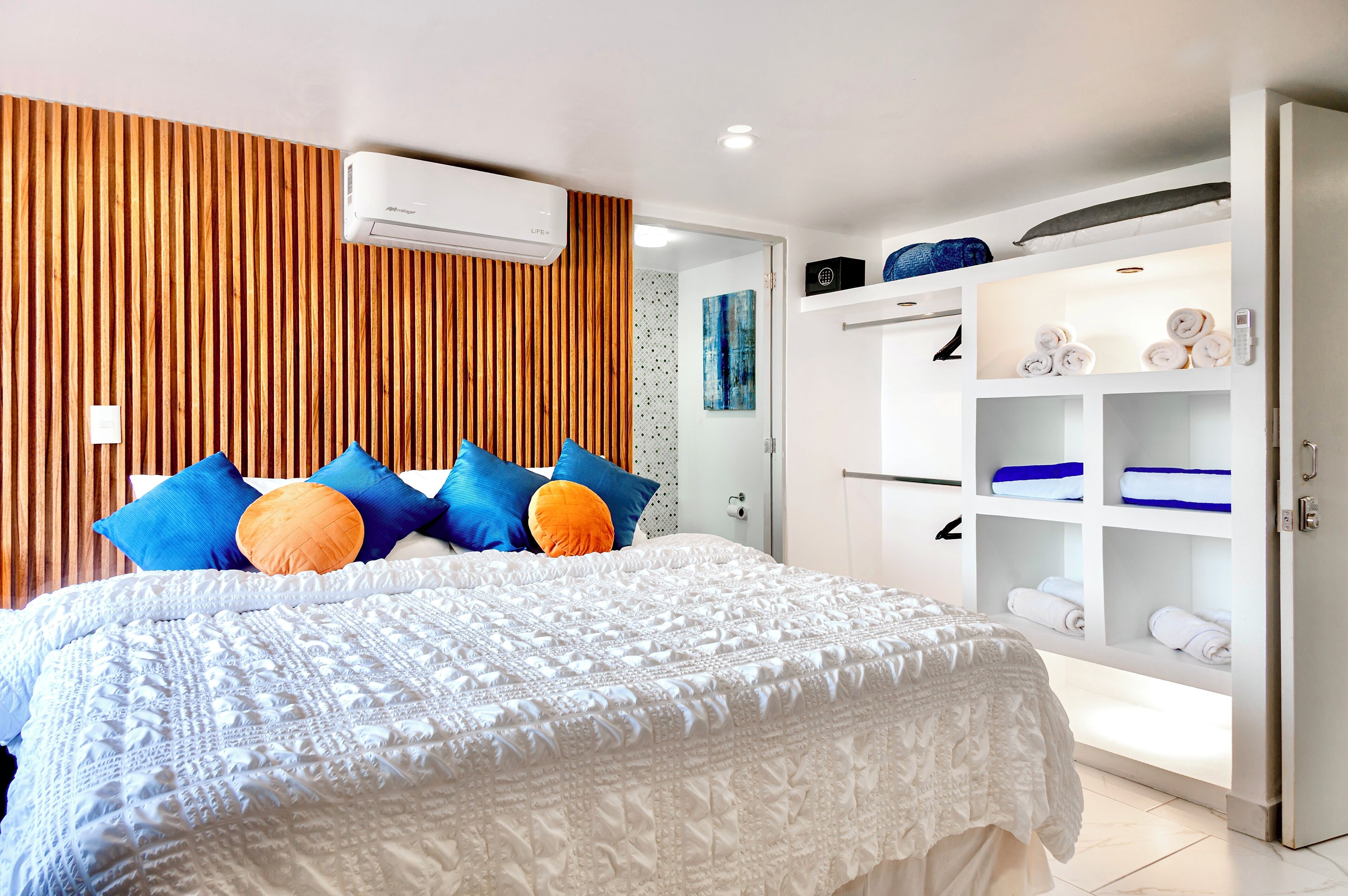 Un dormitorio moderno con una cama grande, un cabecero de listones de madera, un armario blanco con estantes y un ventilador de techo.