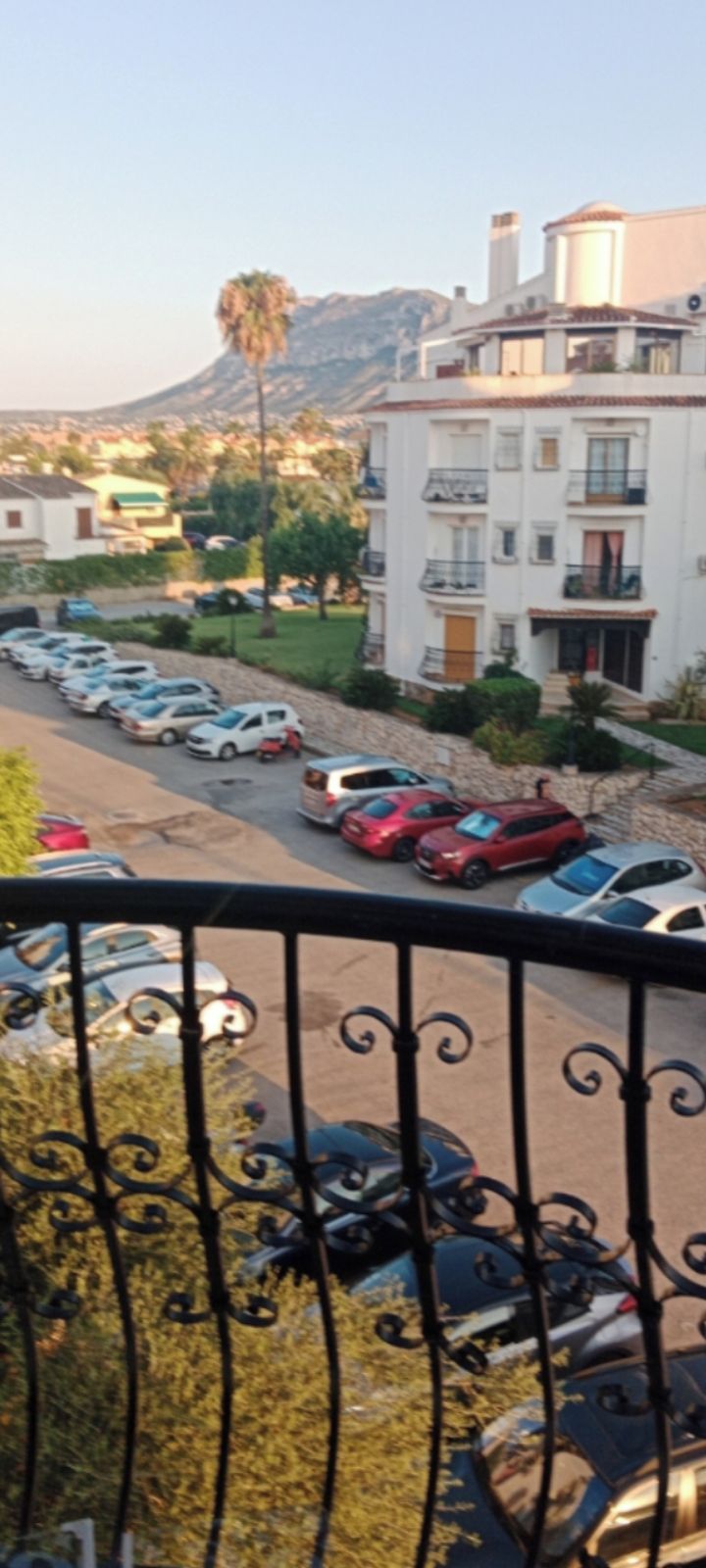 A residential area with a row of parked cars, a white building with balconies, and a mountain in the background.