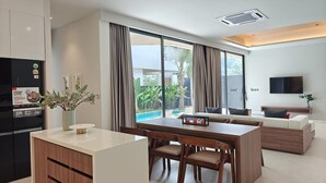 A modern kitchen with a built-in island, a dining area with a table and chairs, a living room with a sofa and a TV, and large windows with curtains. - Aileen Layan Villa 2 Private Pool 4bedroom (Choeng Thale)