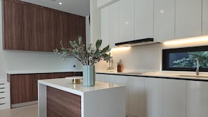 A modern kitchen with white cabinetry, a central island, and a window providing a view of greenery outside. - Aileen Layan Villa 2 Private Pool 4bedroom (Choeng Thale)