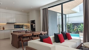 A modern living room with a sofa, dining table, and kitchen in the background. - Aileen Layan Villa 2 Private Pool 4bedroom (Choeng Thale)