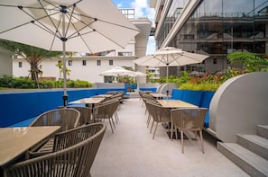 An outdoor patio area with tables and chairs, umbrellas, and a view of a modern building with balconies. - Premier Pool View Studio 348 Beachfront Resort (Choeng Thale)