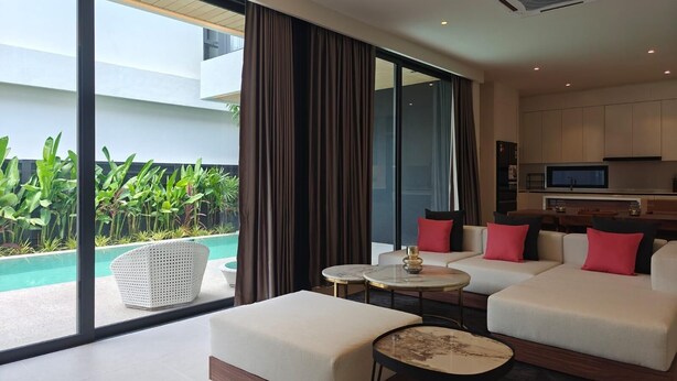 Ein modernes Hotelzimmer mit einer großen Schiebeglasdoor zum Pool, einer Sofagarnitur, einer kleinen Küchenzeile und einem Esstisch.