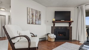 Una sala de estar con chimenea, un televisor de pantalla plana grande montado encima, un sillón cómodo y vistas al exterior desde un balcón.