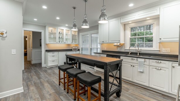Una cocina moderna con muebles blancos, una isla de madera con taburetes y luces colgantes, y un ventanal con vistas a zonas verdes.