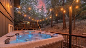 Un jacuzzi en una terraza de madera con luces colgantes, rodeado de árboles.