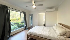 A bedroom with a bed, a ceiling fan, a window with curtains, and a view of greenery outside. - Cute Coomera Home near Theme Parks & Shops (Coomera)