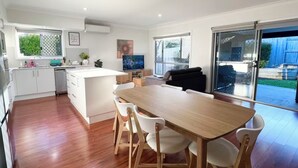 A modern kitchen with white cabinets, a wooden dining table, and a TV. - Cute Coomera Home near Theme Parks & Shops (Coomera)
