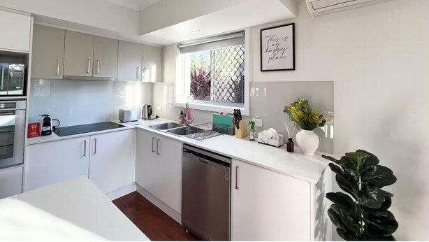 A modern kitchen with white cabinets, a dishwasher, a window with a view, and a framed quote on the wall. - Cute Coomera Home near Theme Parks & Shops (Coomera)