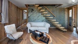 Een moderne woonkamer met een witte bank, een zachte fauteuil en een houten salontafel met een spel. Een houten trap met glazen balustrade leidt naar een andere kamer.