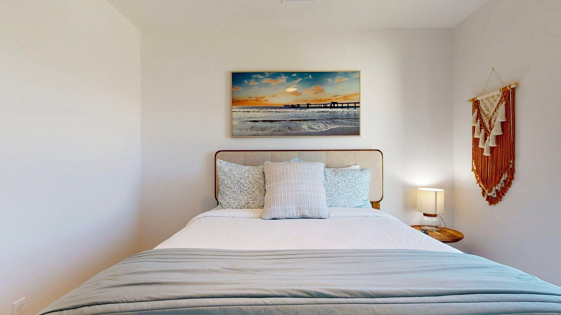 Uma cama bem arrumada com edredom branco e almofadas, um quadro na parede de um pôr do sol sobre um cais, e um tecido pendurado na parede com borlas.