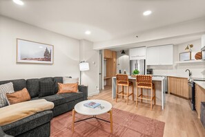 Ein modernes Wohnzimmer mit grauem Sofa, einem runden Couchtisch und einer Küchenzeile mit weißen Schränken und einem Holzessttisch.