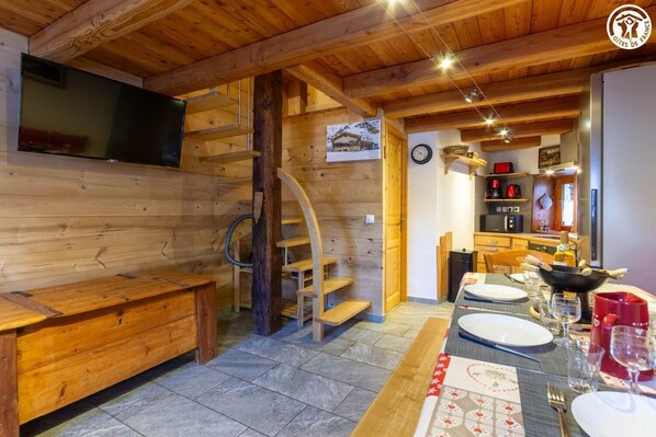 Un intérieur en bois avec un escalier, un coin repas avec une table dressée pour deux personnes, et un espace cuisine équipé d’une cuisinière et d’étagères.