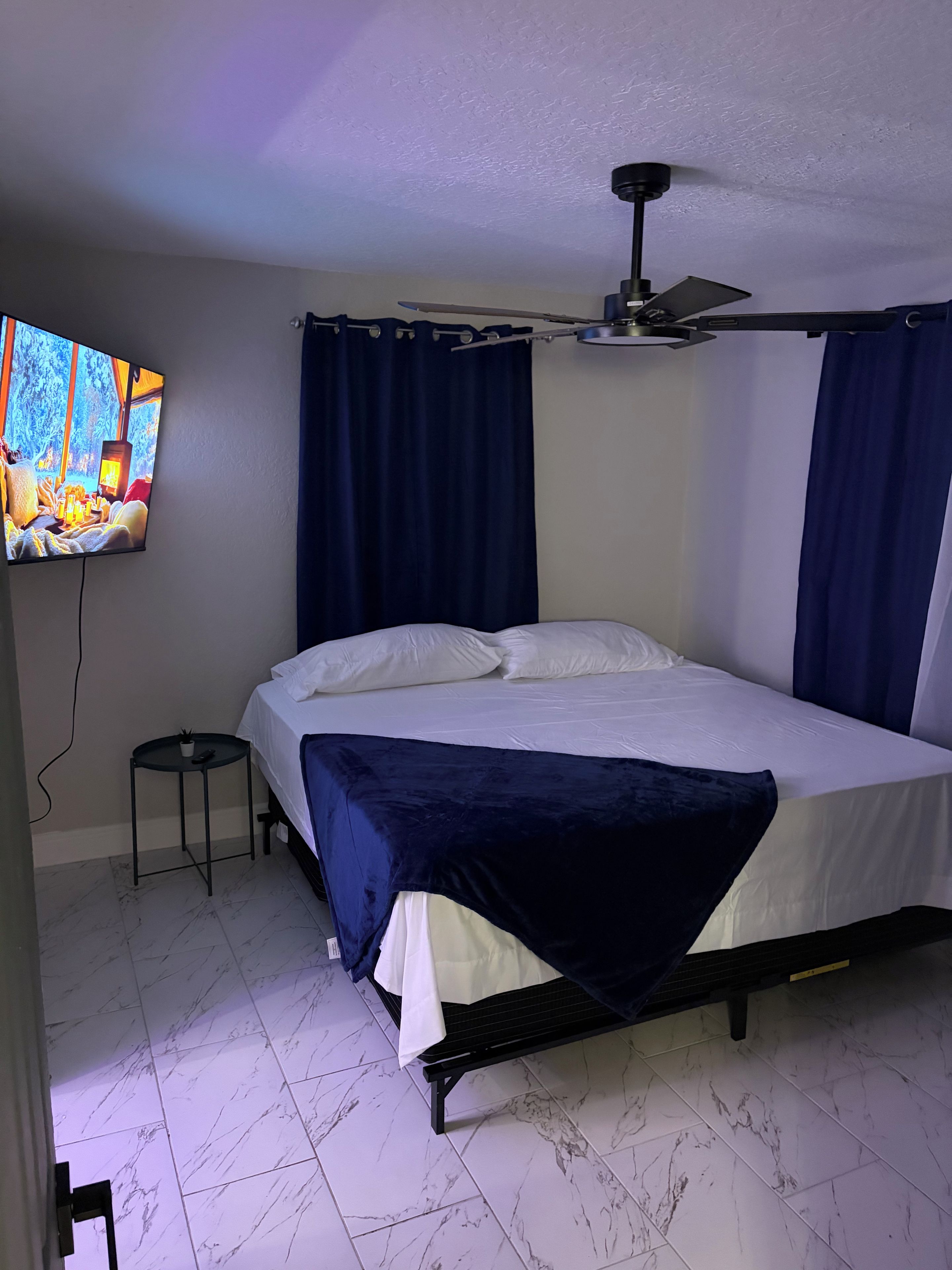 Quarto com cama, ventoinha no teto, televisão fixada na parede e uma pequena mesa com um controle remoto.