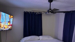 Een slaapkamer met een bed, een plafondventilator, een aan de muur bevestigde televisie en een tafeltje met een afstandsbediening.