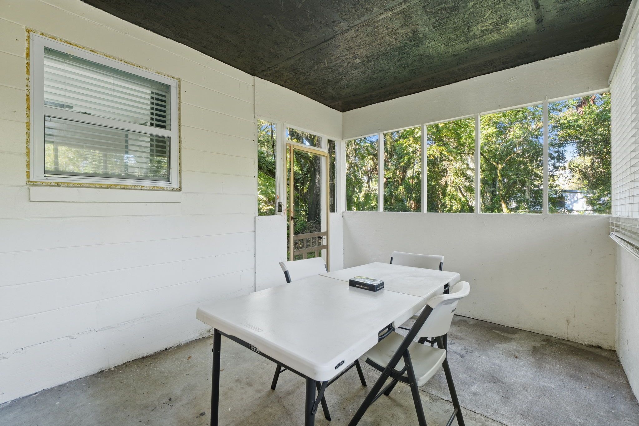 Un pequeño patio cercado con una mesa y sillas blancas, una ventana con persianas y vistas a árboles.