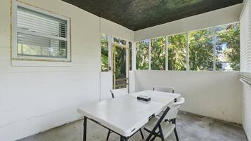 Un pequeño patio cercado con una mesa y sillas blancas, una ventana con persianas y vistas a árboles.