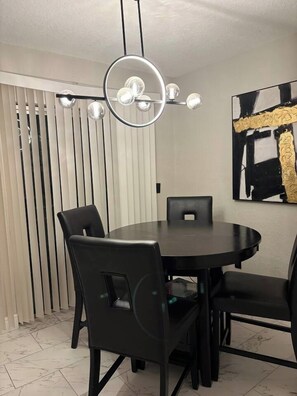 A dining area with a round table and four chairs, a modern chandelier, and a piece of abstract art on the wall.