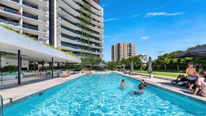 En swimmingpool med folk i vattnet, omgiven av moderna byggnader och ett takförsett sittområde.