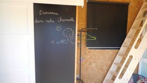 A room with a chalkboard wall displaying 'Bienvenue dans notre Charnotelle' and a simple drawing of a tree and sun. A ladder is leaning against the wall, and a chair with a colorful cushion is visible. - LA CHARMOTTE - Verbiesles (Verbiesles)