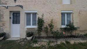 A stone building with a white door, two windows, and a small garden area with plants and a flower bed. - Au petit Café - Châteauvillain (Châteauvillain)