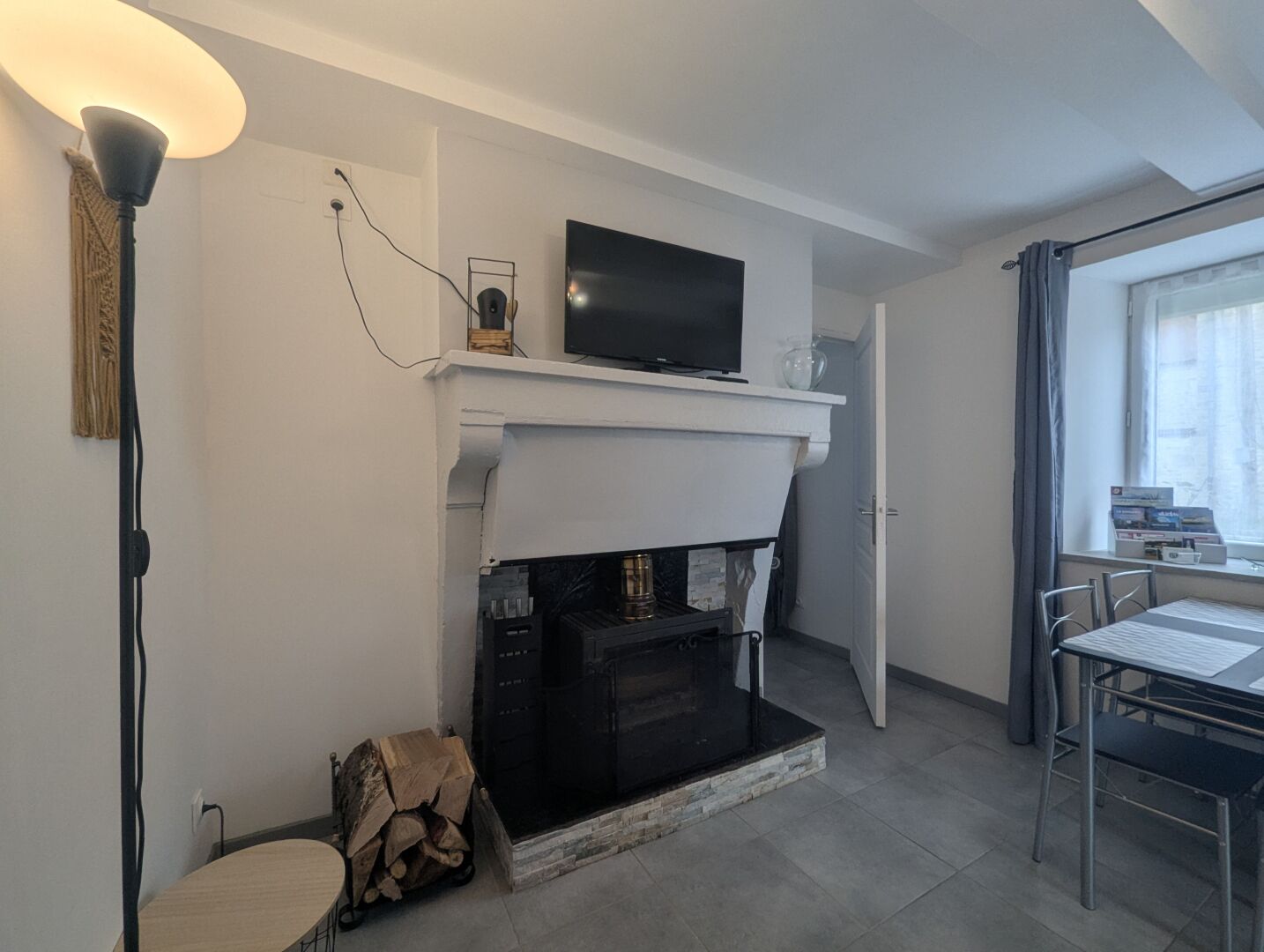 Een moderne woonkamer met een open haard, een flatscreen-televisie erboven, een eettafel met stoelen en een groot raam dat voor natuurlijk licht zorgt.