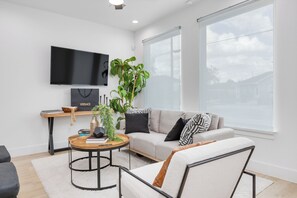 Una sala de estar moderna con un sofá, una mesa de centro, un televisor montado en la pared y un ventanal grande.
