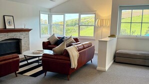 A spacious living room with a fireplace, a leather sofa, a dining table, and large windows offering a view of the countryside.