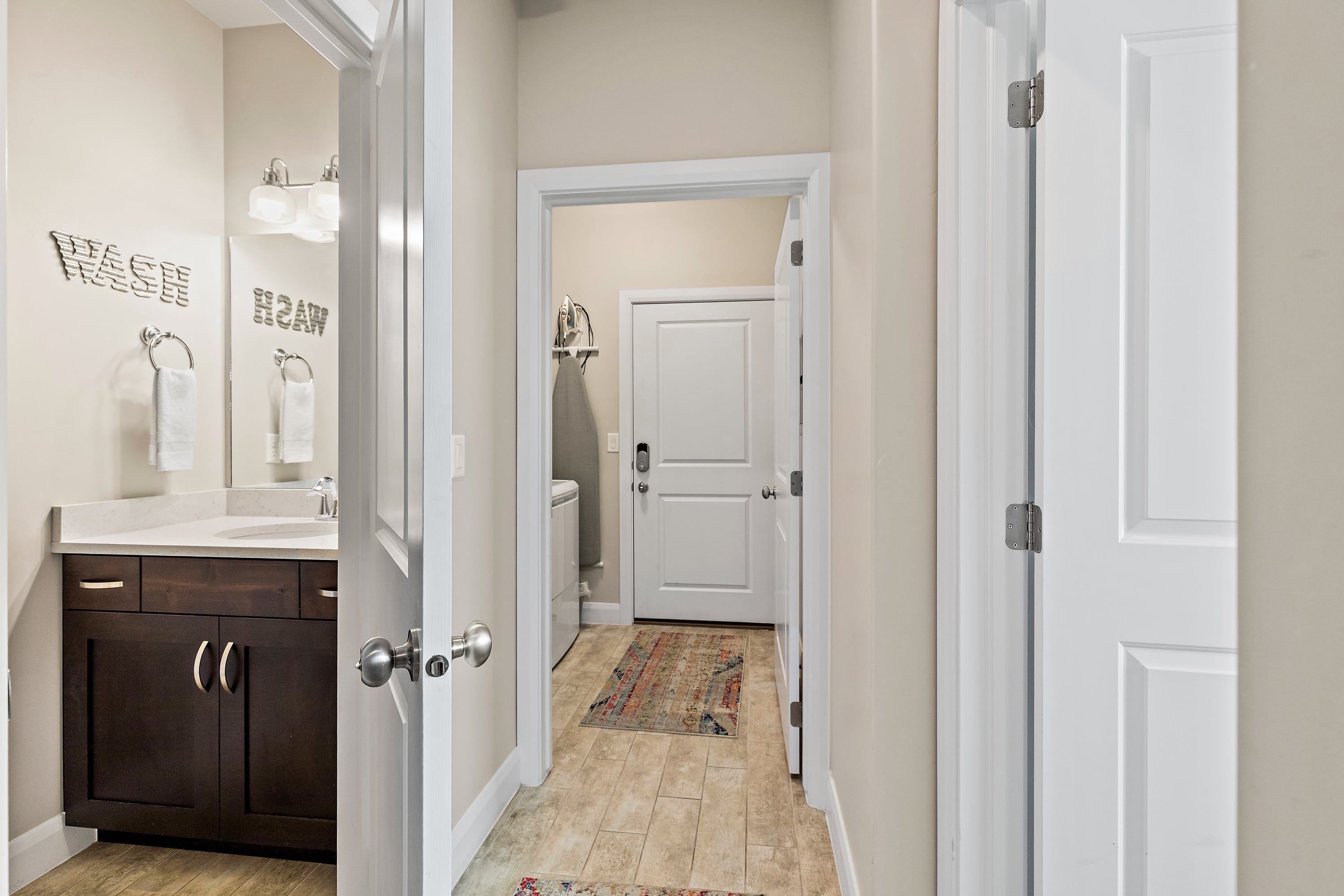 A hallway with a bathroom on the left, featuring a sink, mirror, and towel rack. A door with a towel hanging on it is visible in the background.