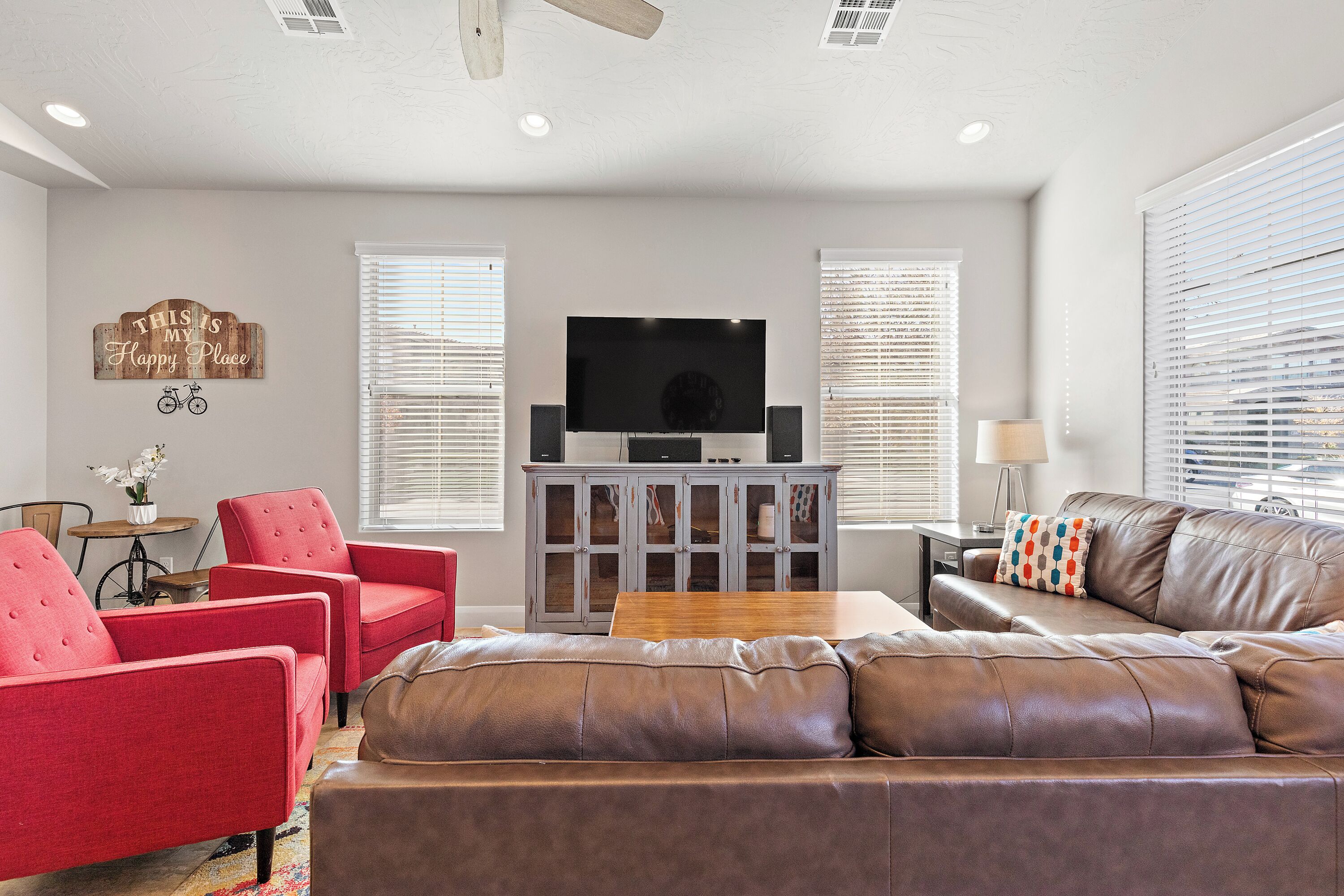 Una sala de estar con un sofá de cuero marrón, sillones rojos, un mueble de madera con televisor de pantalla plana y una mesita.