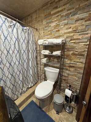 Une salle de bain avec des toilettes, une étagère en métal avec des serviettes, un rideau de douche et une poubelle.