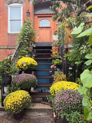 Un edificio de ladrillo con una puerta de madera, una escalera que lleva a la entrada, y varios maceteros con plantas y flores alrededor de los escalones.
