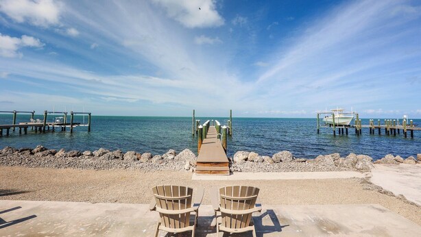 Een strand met een houten pier, twee Adirondack stoelen en een boot aan de kant afgemeerd.