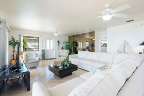 A spacious living room with a white sofa, a coffee table, and a TV. There are ceiling fans, a fireplace, and a large window.