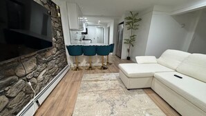 Ein modernes Wohnzimmer mit einer Wand aus Naturstein, einem Flachbildfernseher, einem weißen Sofa und einem Essbereich mit Küche im Hintergrund.