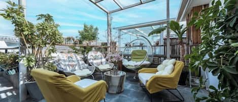 A glass-enclosed patio with seating, plants, and a view of the city.