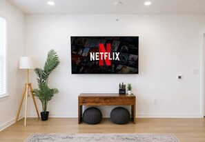 En moderne stue med et stort Netflix-logo på væggen, en træbænk, en potteplante og en gulvlampe.