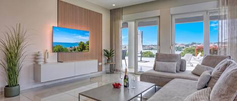 Una sala de estar moderna con una televisión de pantalla plana, un sofá, una mesa de centro y vista al mar a través de amplios ventanales.