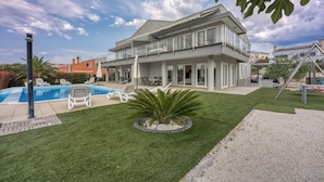 Een modern huis met twee verdiepingen, een zwembad, buitenmeubilair en een verzorgd gazon.