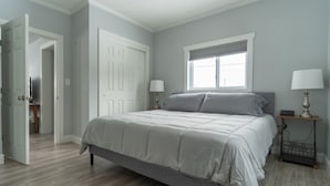 Un dormitorio con una cama, dos mesitas de noche, una ventana con persianas y una puerta que da a otra habitación.