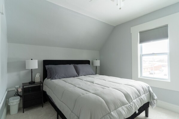 A bedroom with a bed, nightstands, a lamp, a trash bin, and a window with a view.